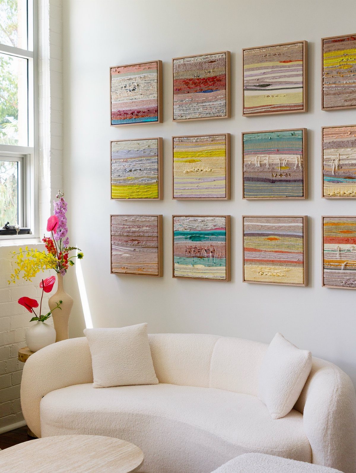 Twelve square woven textile art pieces arranged in a grid above a white sofa