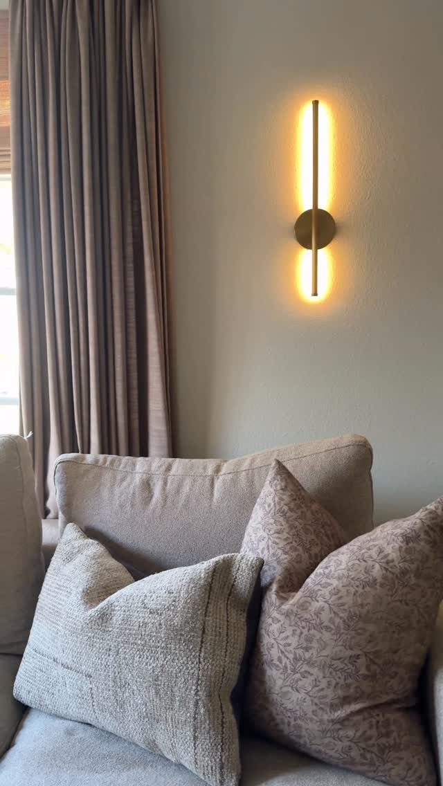 Gold vertical wall sconce glowing on a grey wall above a beige sofa.