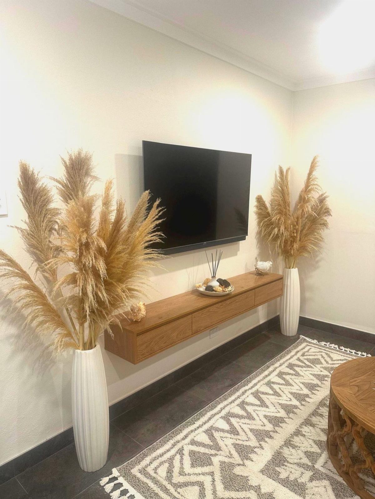 Pampas grass in white vases flanking a television and floating wooden shelf in living room.