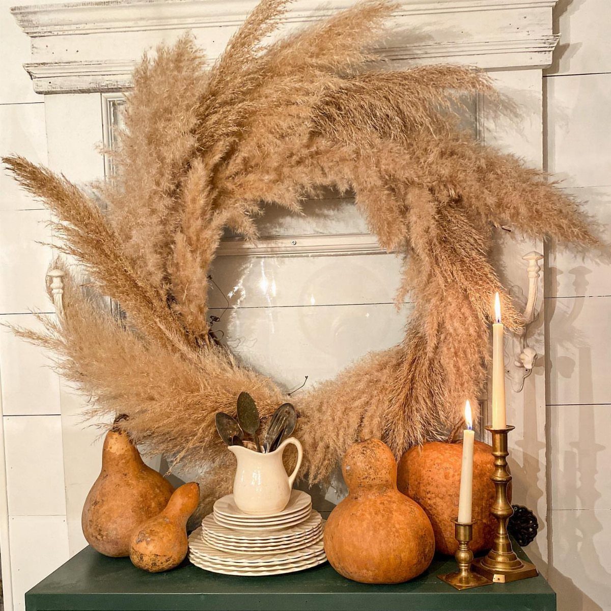 Pampas grass wreath displayed on a mantel with pumpkins and lit candles.
